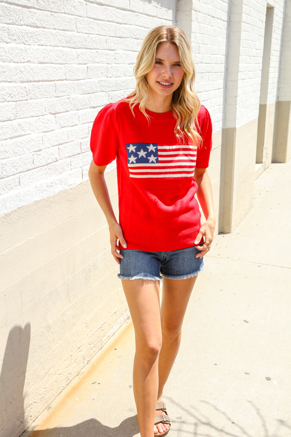 Woman wearing a red t-shirt with an American flag design against a white brick wall.