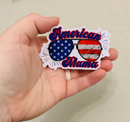 A hand holding a square-shaped sticker magnet with the text 'American Mama' and a design that includes the American flag and sunglasses.