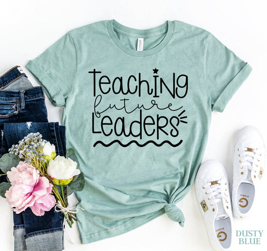 Mint green t-shirt with 'Teaching future leaders' text, paired with jeans, flowers, and sneakers on a white background.